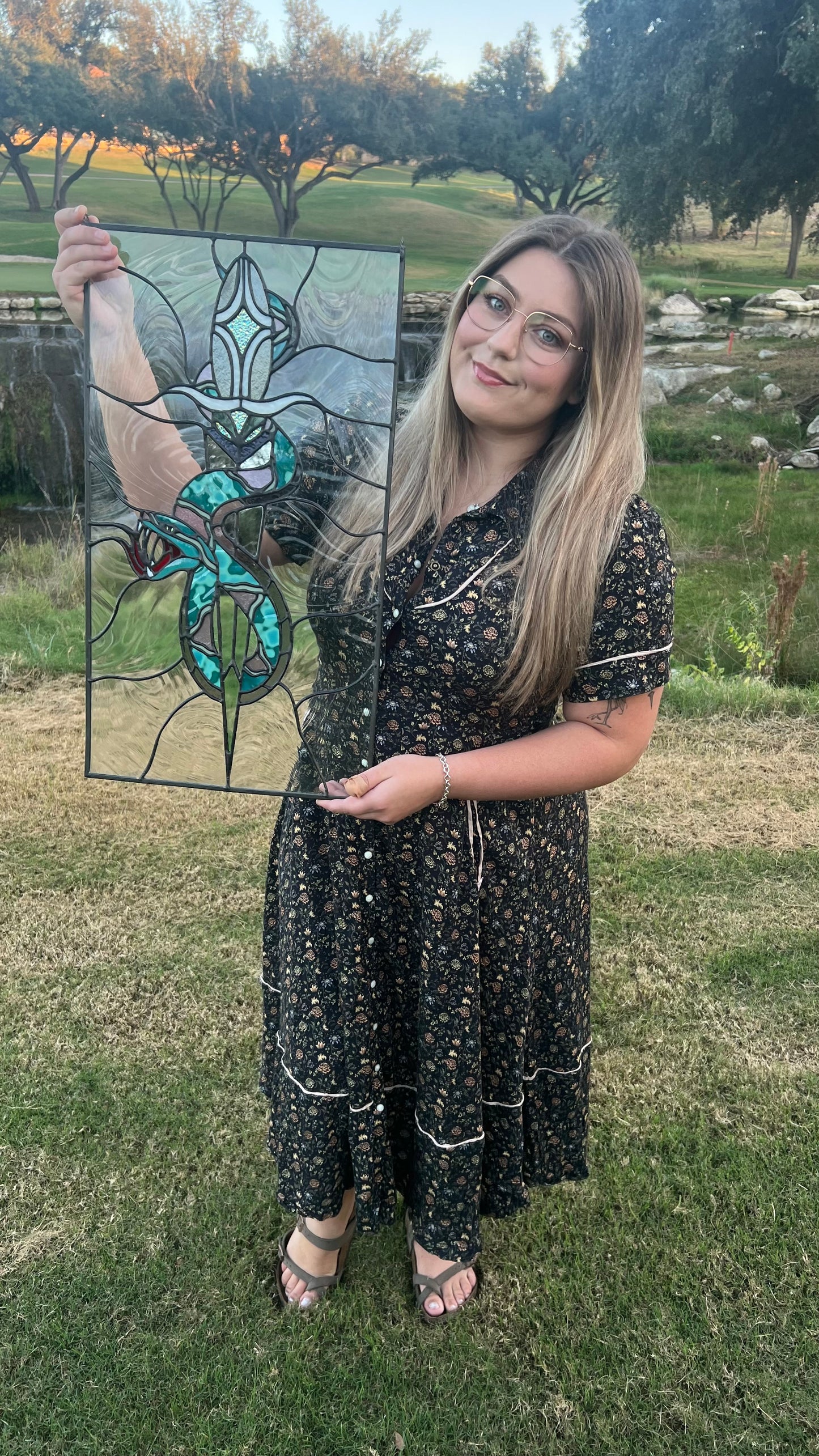 Woman holding a stained glass panel outdoors with greenery in the background