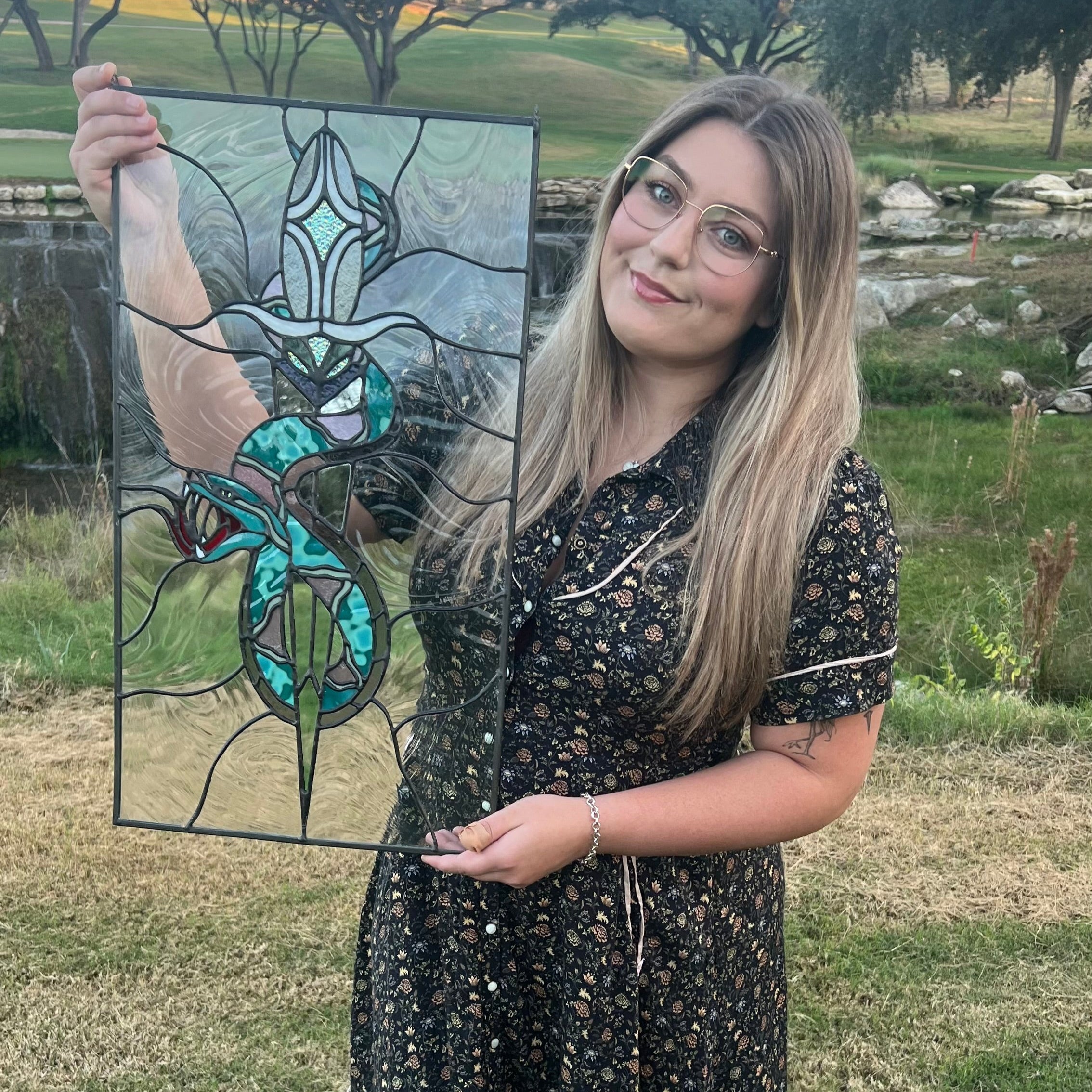 Woman holding a stained glass panel outdoors with greenery in the background
